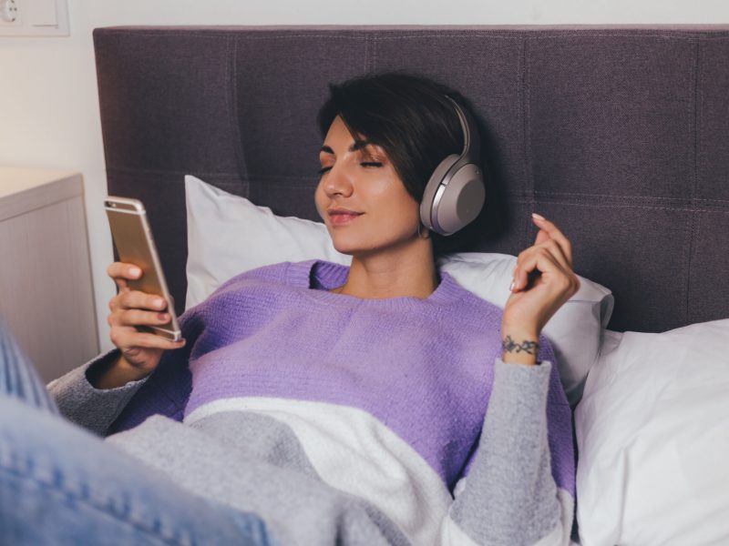 Happy woman at home on comfy bed wearing warm clothes pullover, listen music