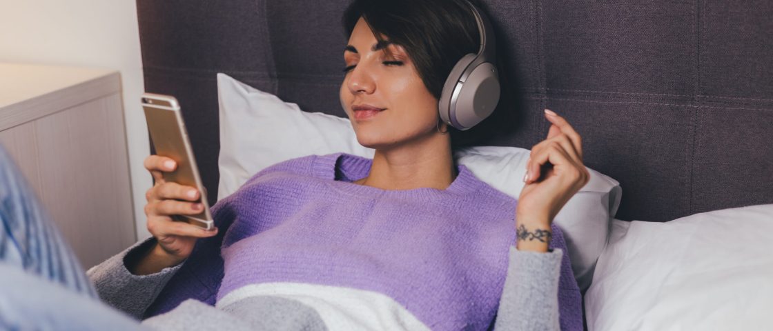 Happy woman at home on comfy bed wearing warm clothes pullover, listen music