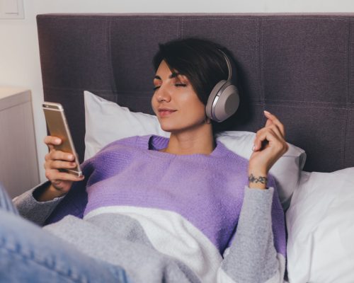 Happy woman at home on comfy bed wearing warm clothes pullover, listen music